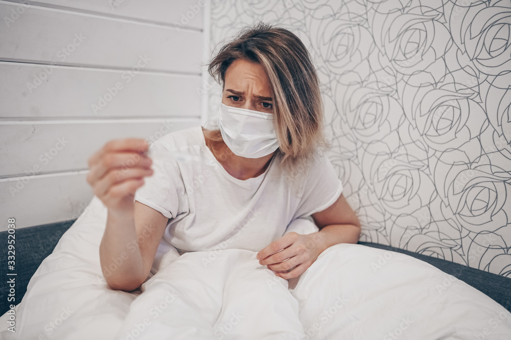 Sick woman lying in bed with thermometer and checking high temperature at home quarantine isolation. Corona virus COVID-19. Infection causes respiratory illness first symptoms cough headache fever Sick woman lying in bed with thermometer and checking high temperature at home quarantine isolation. Corona virus COVID-19. Infection causes respiratory illness first symptoms cough headache fever