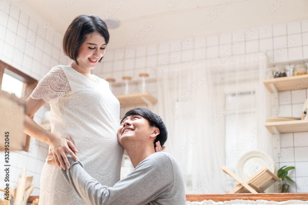 Asian man and pregnancy woman stand in kitchen at home. Father holding mother tummy listen & hear infant inside mom belly. Husband sits and hugs wife. Love and care of pregnant and newborn kid concept Asian man and pregnancy woman stand in kitchen at home. Father holding mother tummy listen & hear infant inside mom belly. Husband sits and hugs wife. Love and care of pregnant and newborn kid concept