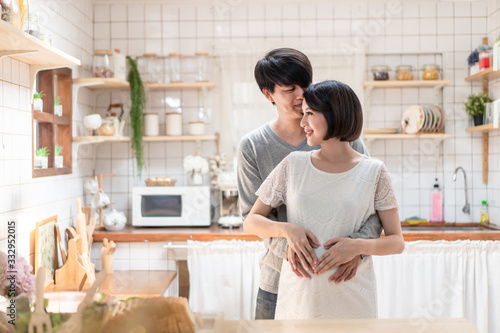 Asian man and pregnancy woman stand in kitchen at home. Father holding mother tummy feeling love expect about future newborn baby. Husband hugs wife from behind. Love and care of pregnant concept.