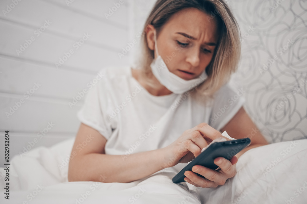 Woman in face protective mask lying in bed during coronavirus isolation home quarantine cleaning phone by hand sanitizer, using cotton wool with alcohol to wipe to avoid contaminating with COVID-19 Woman in face protective mask lying in bed during coronavirus isolation home quarantine cleaning phone by hand sanitizer, using cotton wool with alcohol to wipe to avoid contaminating with COVID-19