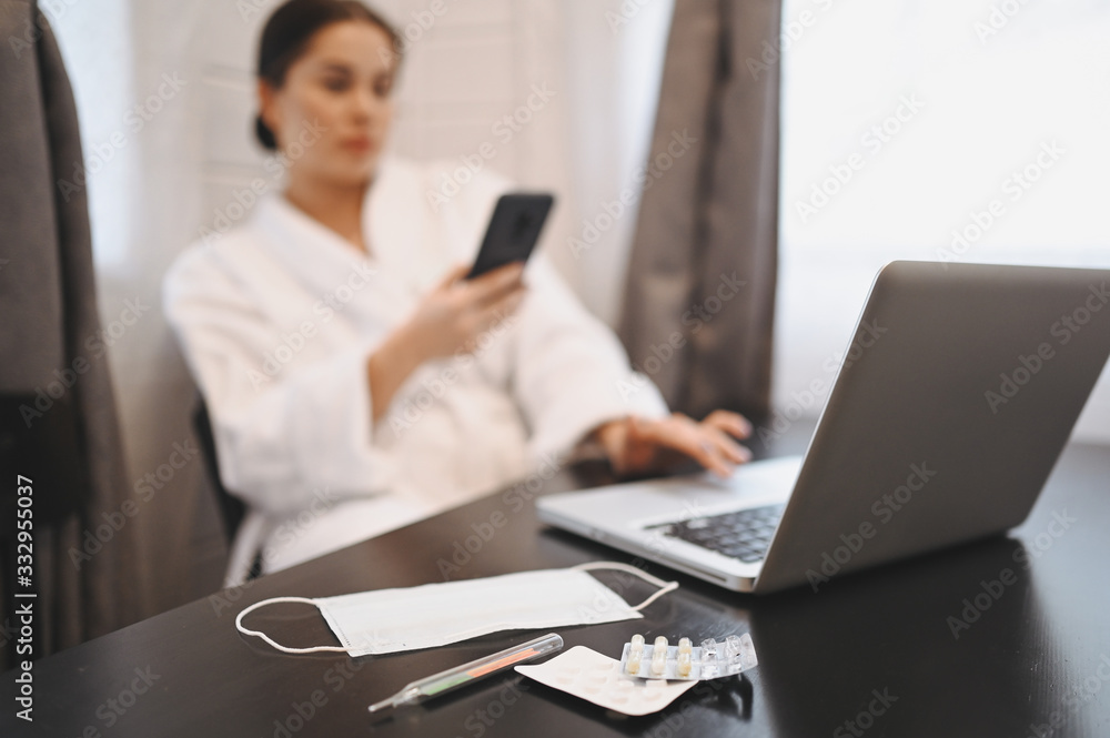 Sick woman working with smartphone and laptop during home quarantine isolation Covid-19 pandemic Corona virus. Distance work from home concept. Thermometer, pills and a medical mask on the table