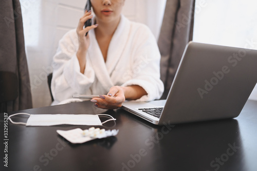 Sick woman speaking smartphone and working on a laptop during home quarantine isolation Covid-19 pandemic Corona virus. Distance work from home concept. Thermometer, pills and a medical mask on table