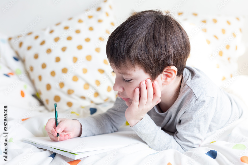 Naklejka premium School Boy on bed and doing school homework at home, writing in notebook. Distance learning online education. Quarantine