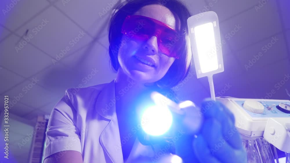 Patient point of view of smiling dentist using curing lights. Beautiful young Caucasian woman in protective eyeglasses and gloves making dental manipulation. Stomatology, medicine.