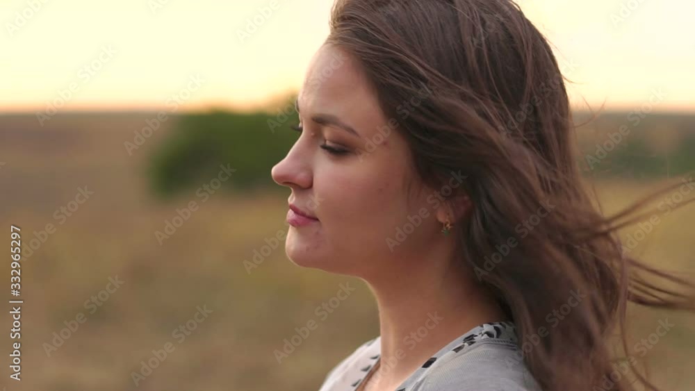 Close-up portrait of a young girl with long dark hair in a field at sunset in a strong wind, she slowly turns her head and her hair flutters in the wind. Close-up portrait of a young girl with long dark hair in a field at sunset in a strong wind, she slowly turns her head and her hair flutters in the wind.