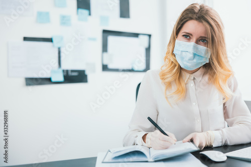 young blond businesswoman manager in medical protection mask and gloves takes notes to her planner working in office during Covid-19 epidemy  virology concept