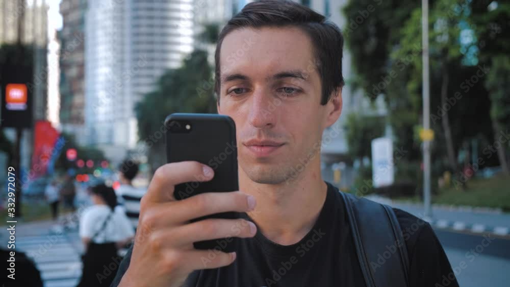 Portrait of tourist man with backpack using smartphone standing outdoors alone smiling at urban city background Portrait of tourist man with backpack using smartphone standing outdoors alone smiling at urban city background