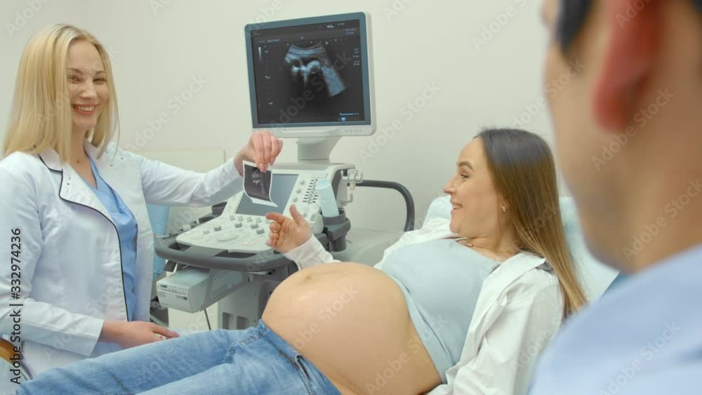 Beautiful pregnant woman lying on examination coach, sharing ultrasound images of baby with husband while female doctor giving consultation. Video in 4K Beautiful pregnant woman lying on examination coach, sharing ultrasound images of baby with husband while female doctor giving consultation. Video in 4K