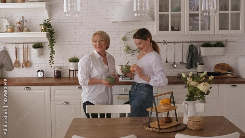 Young woman drinking tea with elderly mother at home. Lovely daughter chatting with her mom at home. An elderly mother treats her adult daughter to pastries in a modern kitchen. Young woman drinking tea with elderly mother at home. Lovely daughter chatting with her mom at home. An elderly mother treats her adult daughter to pastries in a modern kitchen.