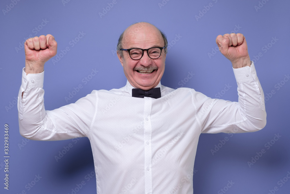 Excited senior handsome man in glasses holding hands up on colored wall Excited senior handsome man in glasses holding hands up on colored wall
