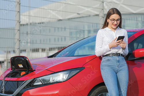 Woman stands with phone near her red electric car and waits when vehicle will charged.