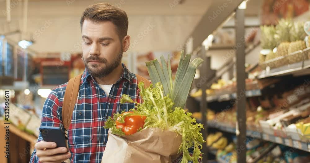 Caucasian young male customer holding packet with fresh vegetables and fruit and walking supermarket isle. Handsome man having shopping list on his smartphone while buying in shop. Caucasian young male customer holding packet with fresh vegetables and fruit and walking supermarket isle. Handsome man having shopping list on his smartphone while buying in shop.