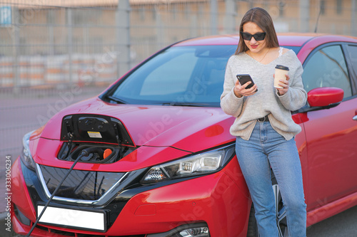 Woman stands with phone near her electric car and waits when vehicle will charged. Charging of electric car © uflypro