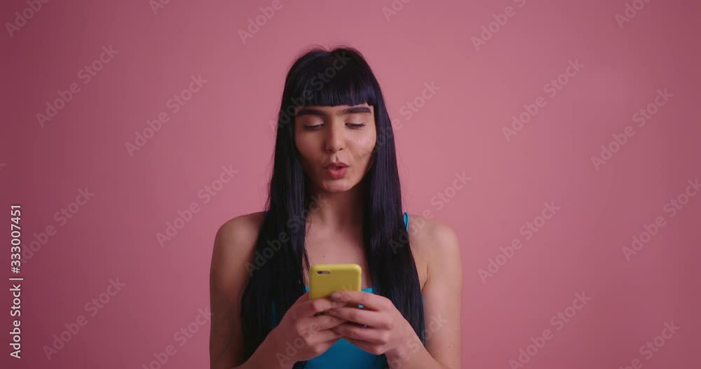 A pretty smiling brunette woman in blue shirt using smartphone isolated on pink background A pretty smiling brunette woman in blue shirt using smartphone isolated on pink background