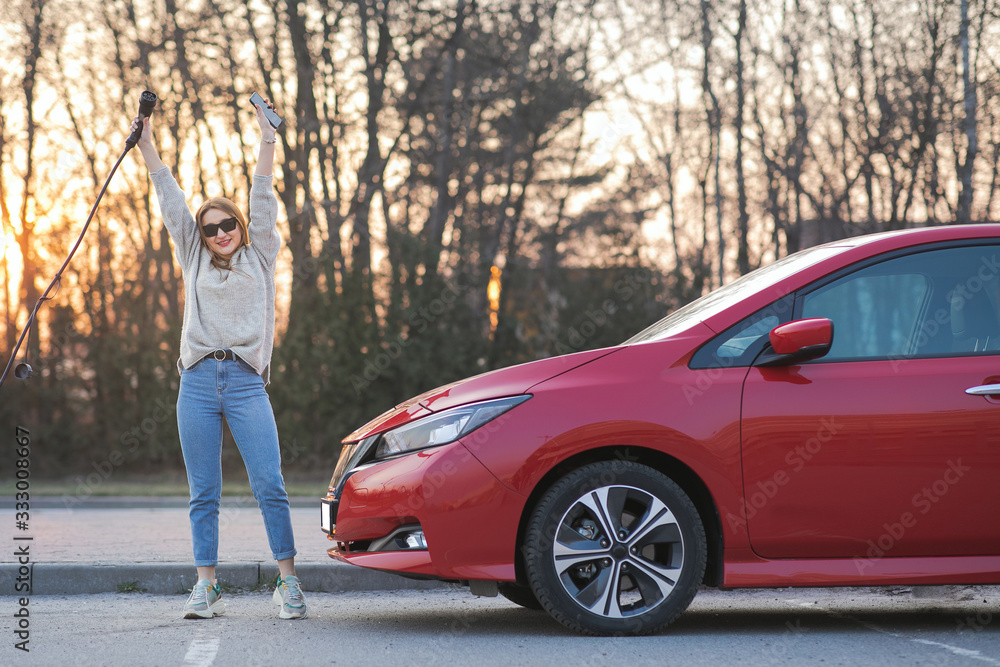 Young girl looking at camera having fun and holding in hand power cable supply standing near electric car at charging station Young girl looking at camera having fun and holding in hand power cable supply standing near electric car at charging station