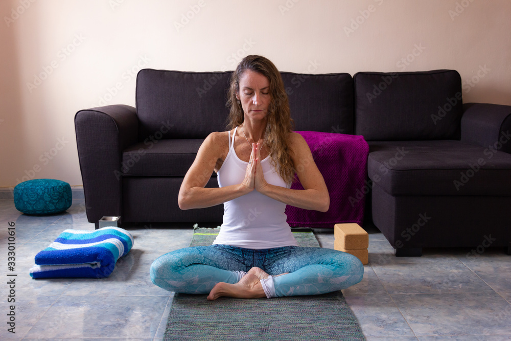 Yoga teacher with hands together in anjali mudra praying pose on chest in living room. Middle aged woman sitting in padmasana with props around her. Working from home, female yogi practice concepts Yoga teacher with hands together in anjali mudra praying pose on chest in living room. Middle aged woman sitting in padmasana with props around her. Working from home, female yogi practice concepts