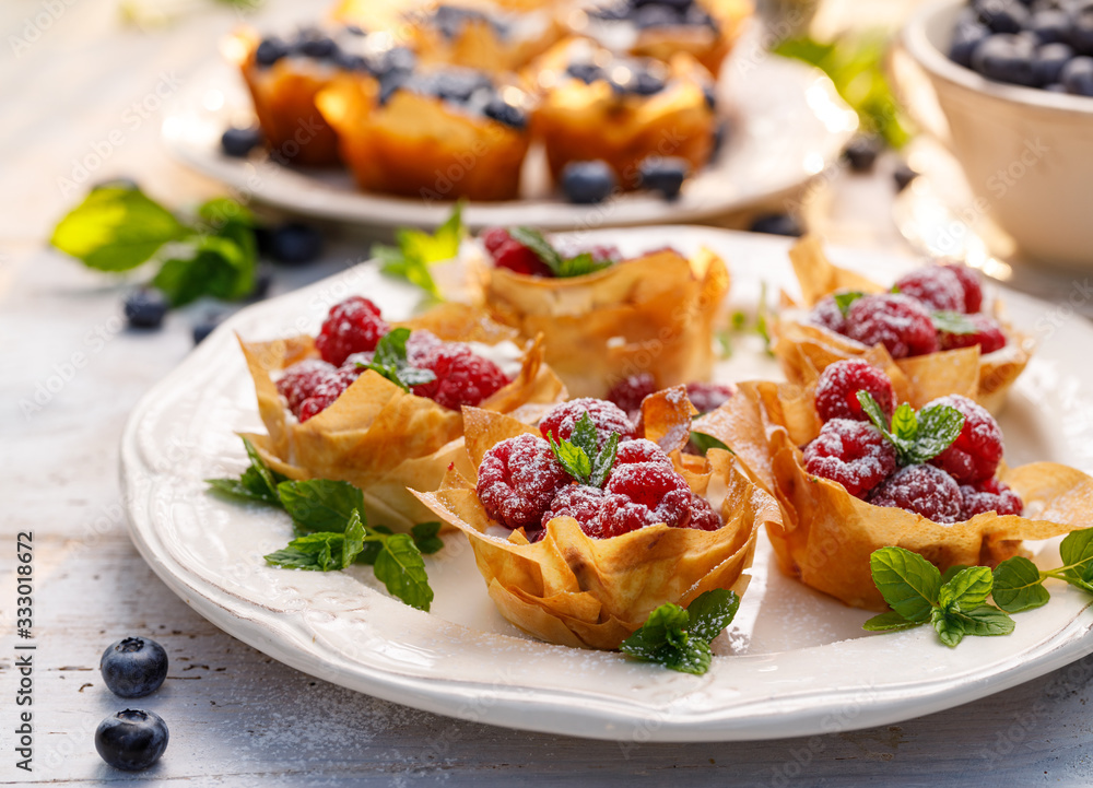 Phyllo cups with Mascarpone cheese filling topped with fresh raspberries and mint on a white plate, close up. Delicious filo pastry dessert