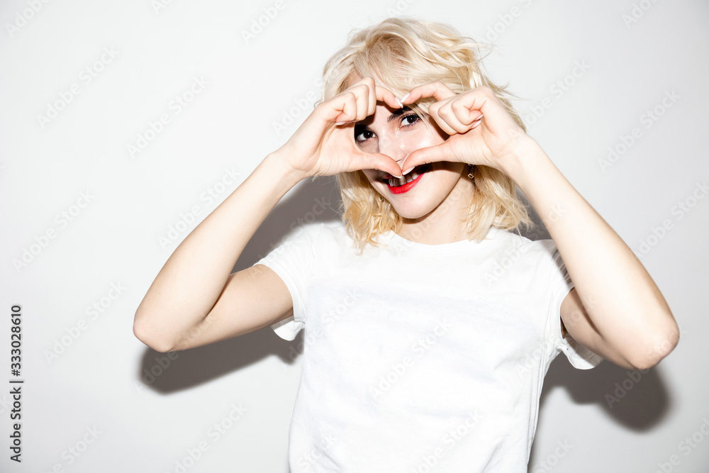 lovely young blonde girl in a white t-shirt on a white background shows a symbol of love, studio fashion photo with copy space lovely young blonde girl in a white t-shirt on a white background shows a symbol of love, studio fashion photo with copy space