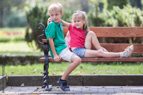 Portrait of two pretty cute children boy and girl having fun time on a bench in summer park outdoors. © bilanol