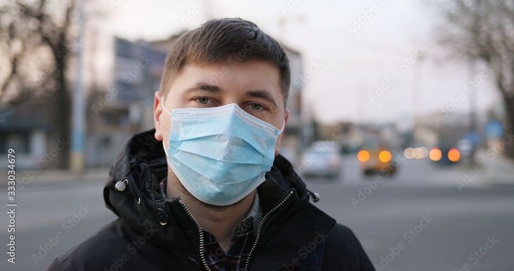 Portrait of Caucasian upset ill man in medical mask standing at street and looking to camera. Close up of unhealthy guy wearing protection from coronavirus in city. Pandemic concept. Infected person.