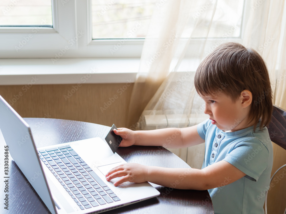 Curious toddler boy explores the laptop and presses buttons on computer keyboard. Kid holds credit card in hand. Puzzled child. Curious toddler boy explores the laptop and presses buttons on computer keyboard. Kid holds credit card in hand. Puzzled child.