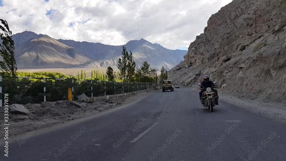 Parallax shot of two people riding a motorcycle on the scenic Leh-Manali Highway between the rocky slopes of Himalaya mountains passing cars and bikes in Ladakh, India Parallax shot of two people riding a motorcycle on the scenic Leh-Manali Highway between the rocky slopes of Himalaya mountains passing cars and bikes in Ladakh, India