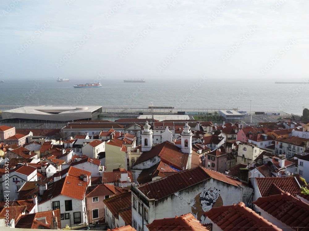 View of the Lisbon area from above, with white houses and red roofs and the river Tejo in the background.