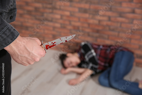 Man with bloody knife and his victim on floor indoors, closeup. Dangerous criminal