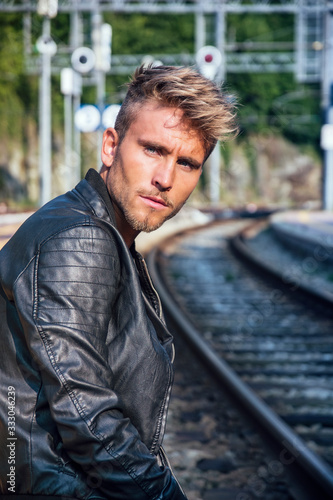 Attractive blond young man standing by railroad tracks, wearing black leather jacket and looking at camera © starsstudio