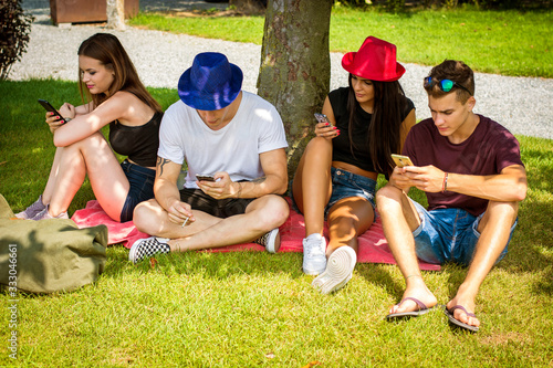 Group of young people sitting under tree, using their smartphones and communicating with each other or absorbed with them. Concepts of lack of communication, self-absorbtion, overuse of technology