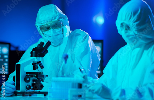 Scientists in protection suits and masks working in research lab using laboratory equipment: microscopes, test tubes. Medicine, coronavirus 2019-ncov infection and vaccine discovery.