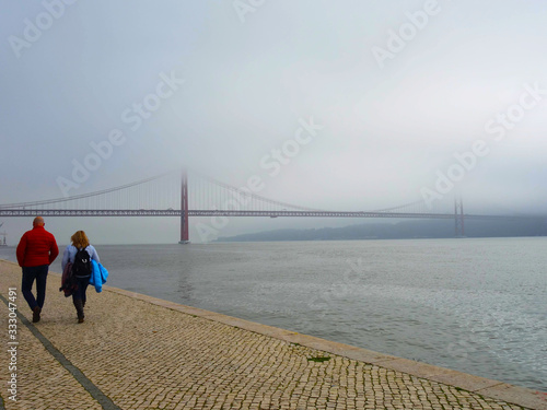 Lisbon, Portugal-23 December 2019: people walking along Lisbon's waterfront, in the background the 25 April bridge crossing the river Tejo on a foggy day.