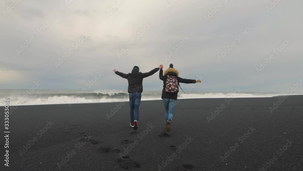 Young hipster couple running on volcanic black sand beach in Iceland and having fun at sunset, slow motion. Reynisfjara Beach, Reynisdrangar troll toes mountains, Vik. Iceland in winter Young hipster couple running on volcanic black sand beach in Iceland and having fun at sunset, slow motion. Reynisfjara Beach, Reynisdrangar troll toes mountains, Vik. Iceland in winter