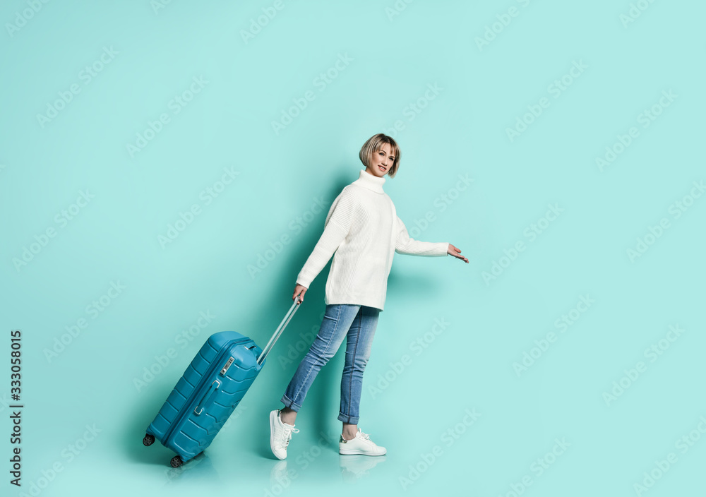 Blonde girl in white sweater, jeans and sneakers. She is smiling posing with suitcase luggage standing sideways on blue Blonde girl in white sweater, jeans and sneakers. She is smiling posing with suitcase luggage standing sideways on blue