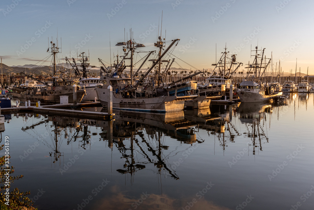 Fishing and squid boats moored in the safety of the harbor as the tall masts are reflected in the ocean water surface.