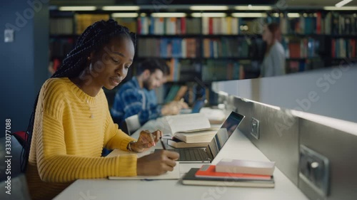 University Library: Gifted Black Girl uses Laptop, Writes Notes for the Paper, Essay, Study for Class Assignment. Diverse Multi-Ethnic Group of Students Learning, Studying for Exams, Talk in College photo