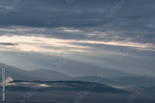 clouds and rays of dawn in mountain valley, soft light in early morning, meditation in nature. Gentle hills in bluish haze, silhouettes of mountains