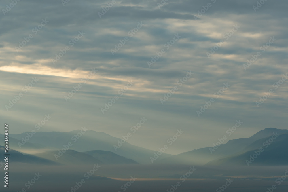 Fototapeta premium clouds and rays of dawn in mountain valley, soft light in early morning, meditation in nature. Gentle hills in bluish haze, silhouettes of mountains