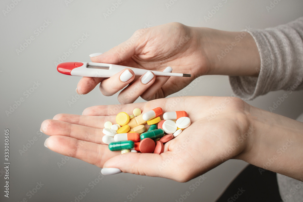 A handful of various pills with a digital thermometer in a woman's hands. Medications against the COVID-19 and other conditions and diseases on the grey background. Medicine. A handful of various pills with a digital thermometer in a woman's hands. Medications against the COVID-19 and other conditions and diseases on the grey background. Medicine.