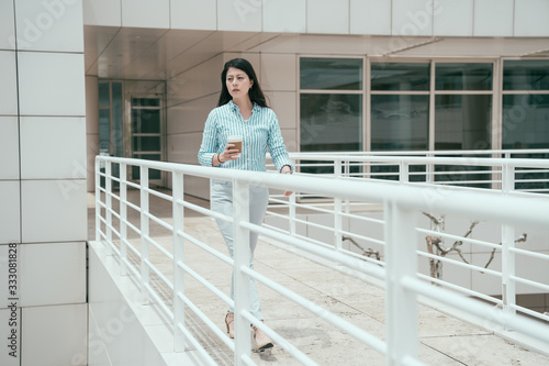full length confident asian korean businesswoman walking with coffee on modern bridge at business center. young elegant office lady with hot drink in disposable paper cup passing on sky walkway.