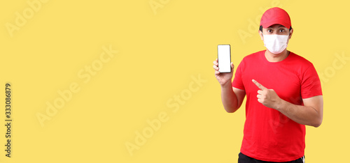handsome Asian man in red cap,wearing a mask is sick Pointing finger isolated on yellow background in studio With copy space. holding mobile phone with blank white empty screen.