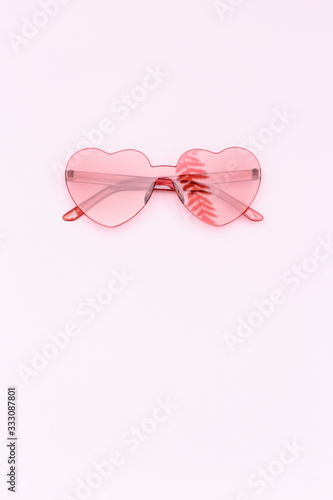 Minimal style fashion photography with heart shaped glasses on pink background with copy space. Pink modern sunglasses and reflection of palm leaf. Trendly summer concept.