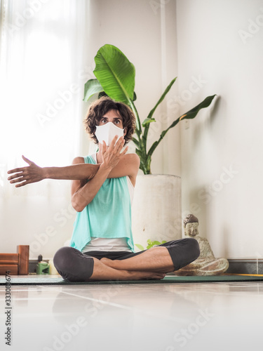 woman in crossed legged position stretching arm and wearing a medical face mask to protect from corona virus covid-19 on a yoga mat in a studio with cushions, buddha and palm tree in background