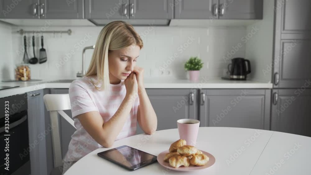 Sad woman with tablet, cup of coffee and croissants on her kitchen Sad woman with tablet, cup of coffee and croissants on her kitchen