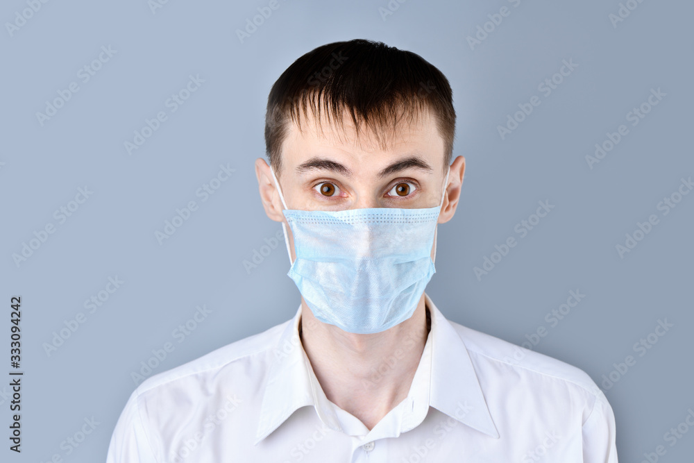 Man in a medical mask. Surprised, scared man in a medical mask in a white shirt looking at camera , on a gray background. Chinese Coronavirus , Covid-19 Man in a medical mask. Surprised, scared man in a medical mask in a white shirt looking at camera , on a gray background. Chinese Coronavirus , Covid-19