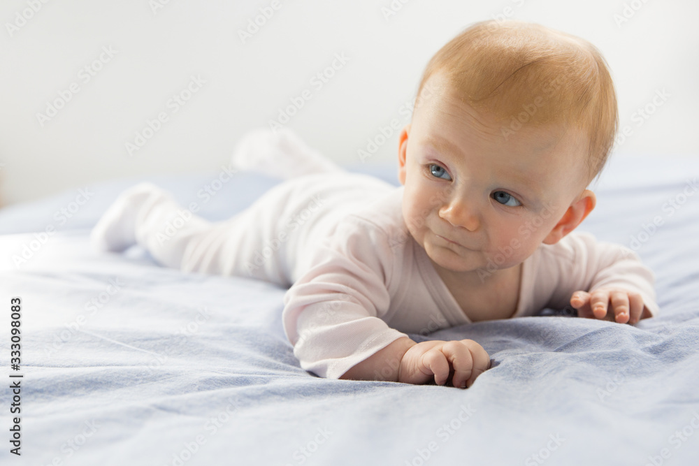 Positive funny blue eyed baby girl crawling on bed sheet. Six month child lying on belly at home. Childhood or baby care concept Positive funny blue eyed baby girl crawling on bed sheet. Six month child lying on belly at home. Childhood or baby care concept