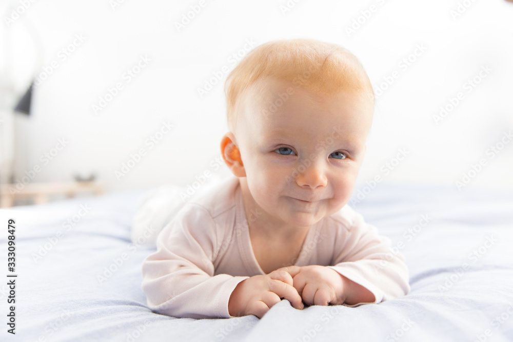 Joyful smiling red haired baby girl crawling on bed sheet and looking at camera. Six month child lying on belly in bedroom. Childhood or baby care concept Joyful smiling red haired baby girl crawling on bed sheet and looking at camera. Six month child lying on belly in bedroom. Childhood or baby care concept