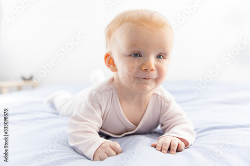 Funny joyful red haired baby girl crawling on bed linen. Six month child lying on belly in bedroom. Childhood or baby care concept