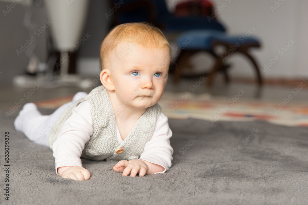 Beautiful red haired baby girl crawling on grey carpet at home. Six month child lying on belly on floor. Childhood or baby care concept Beautiful red haired baby girl crawling on grey carpet at home. Six month child lying on belly on floor. Childhood or baby care concept