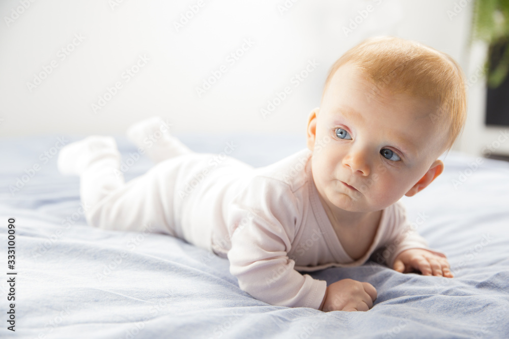 Serious pensive cute baby girl crawling in bedroom. Six month child lying on belly at home. Childhood or baby care concept Serious pensive cute baby girl crawling in bedroom. Six month child lying on belly at home. Childhood or baby care concept